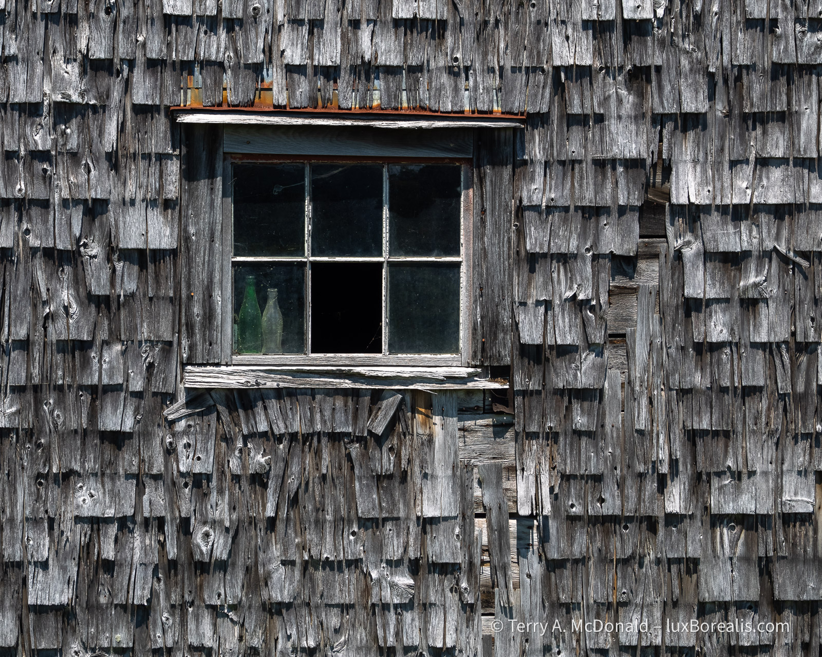 Passing the Test
A wall of decaying grey cedar shakes surrounds an old window with some green and clear bottles inside on the window sill.