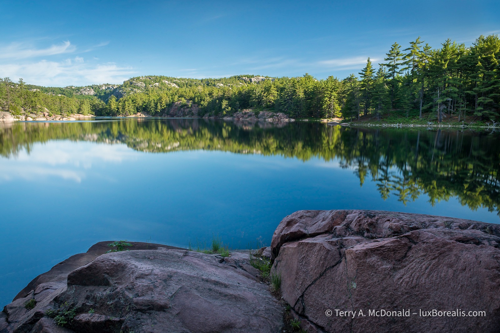 Dawn, A.Y. Jackson Lake, Killarney
A clear, blue sky dawns over A.Y. Jackson Lake, named for a member of the Group of Seven, with pink granite in the foreground and a rocky shoreline forested with coniferous trees. ©Terry A. McDonald – luxBorealis.com