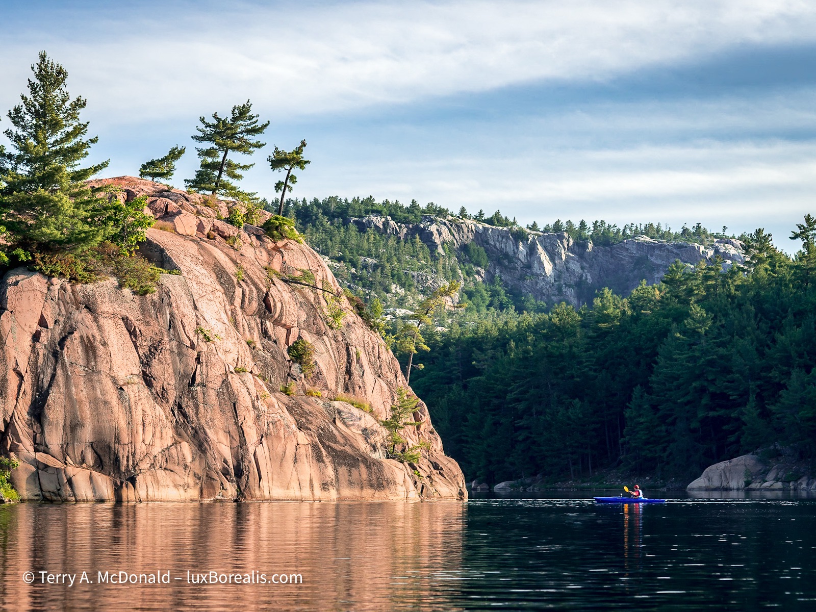 Morning Paddle, George Lake, Killarney
A lone paddler in a blue kayak looks up at the pink glow coming from the George Lake monolith of granite with the white quartzite LaCloche Range behind. ©Terry A. McDonald – luxBorealis.com
