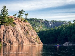 Morning Paddle, George Lake, Killarney – A lone paddler in a blue kayak looks up at the massive, [ink granite George Lake monolith, with the white quartzite hills of the LaCloche Range behind. ©Terry A. McDonald – luxBorealis.com