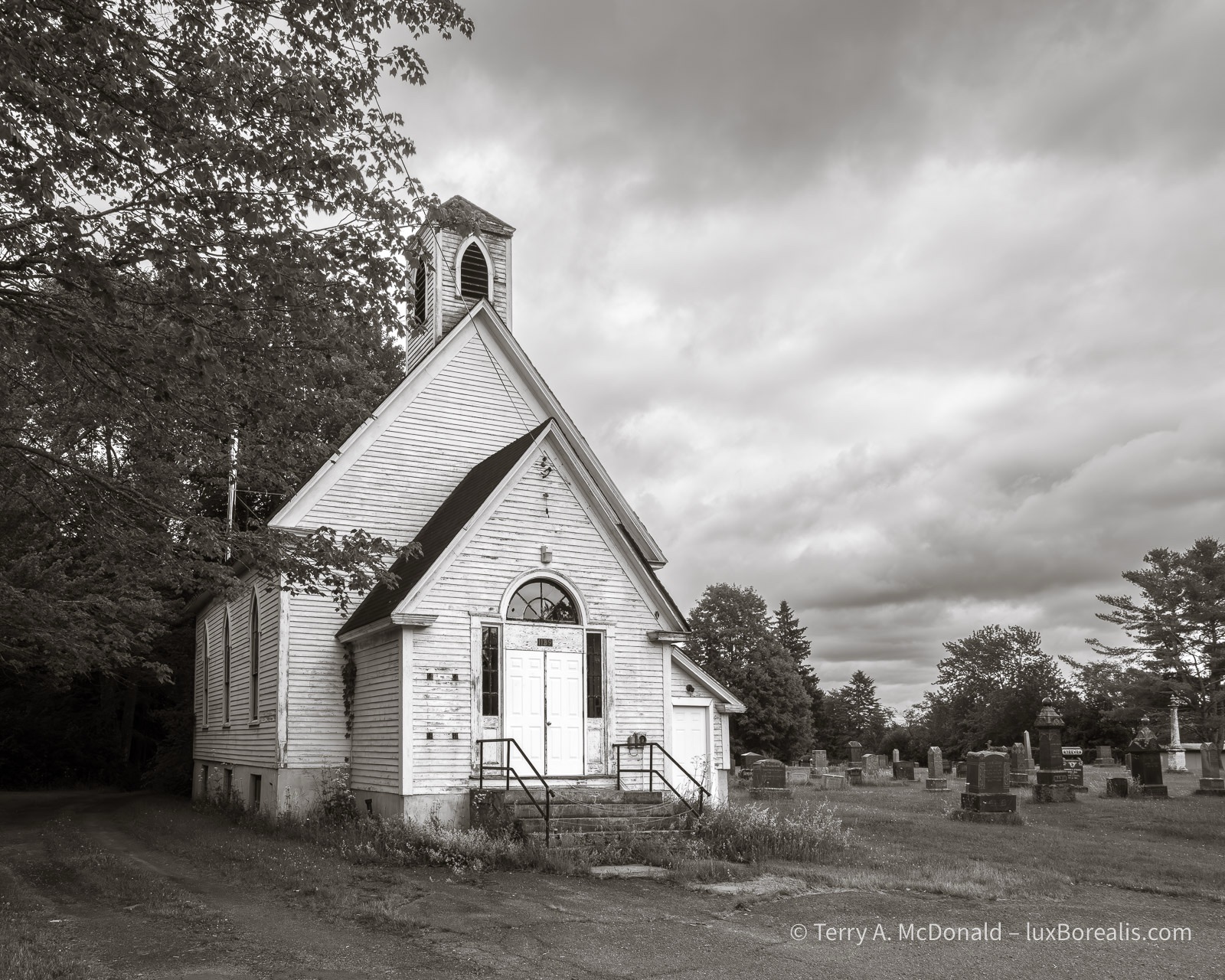 Forlorn
In this black-and-white photograph, a worn, tired, dilapidated church, painted white, stands against its graveyard under a menacing sky of clouds.