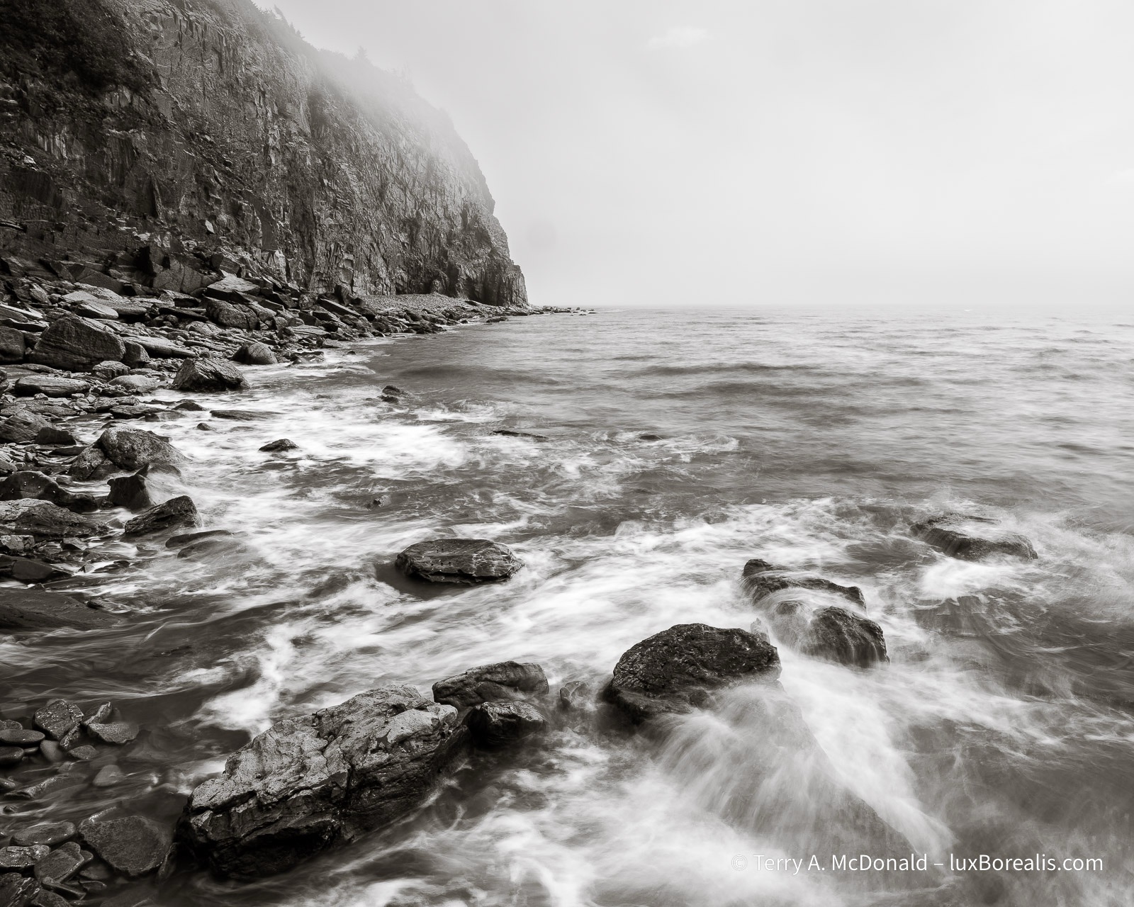 W=In the black-and-white photograph, wave wash is coming ashore over the boulders and cobbles at Cape Enrage, with the cliffs and fog in the background.