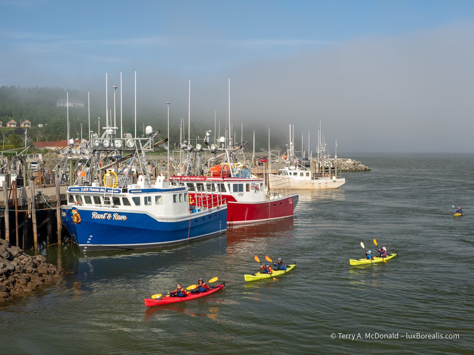 Alma Harbour, High Tide
In bright sunshine, kayakers paddle past brightly coloured fishing boats are their way into the harbour, with a bank of fog obscuring the horizon.