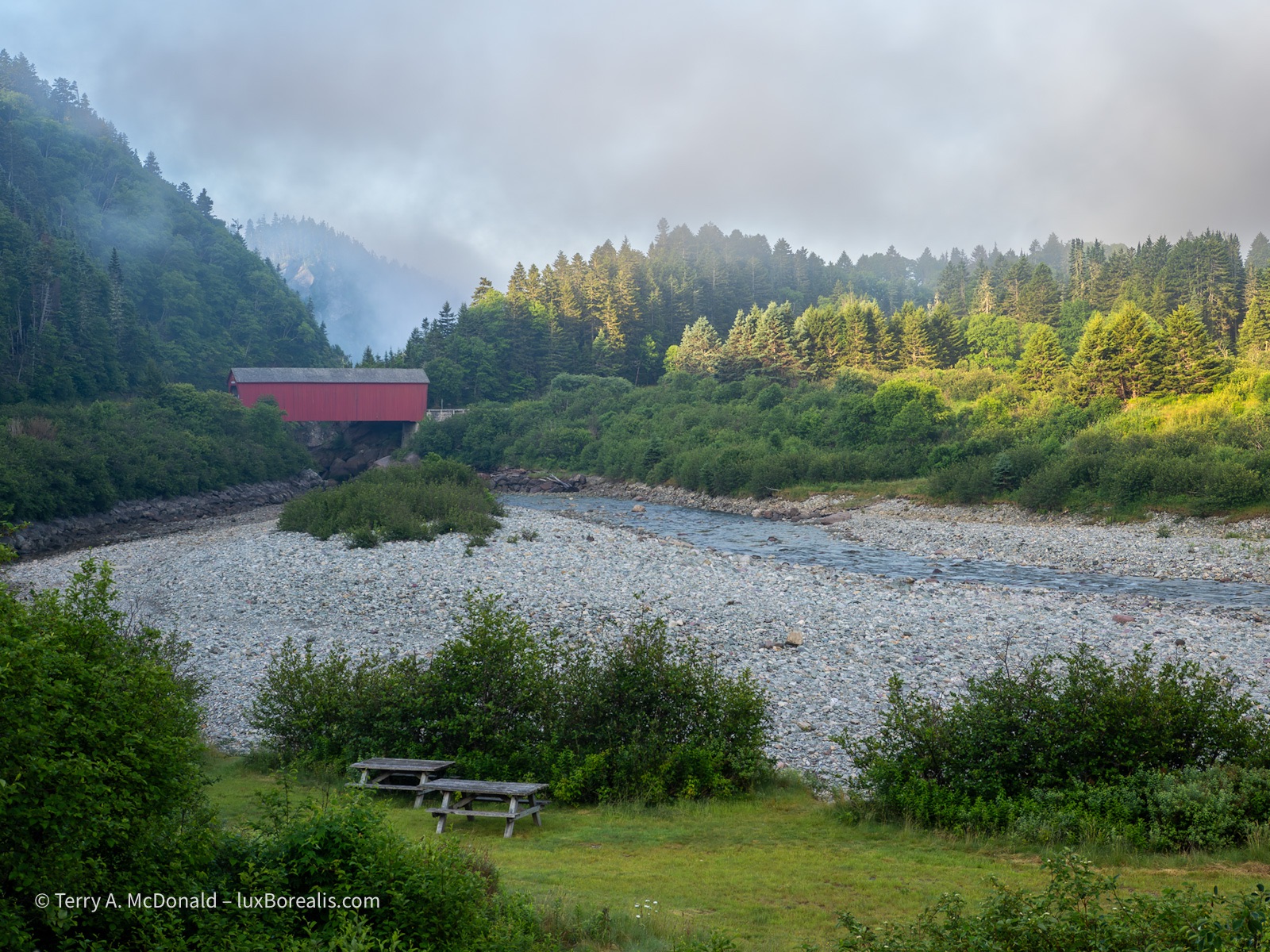 Morning, Wolfe Point Covered Bridge
The bright red Wolfe Point covered bridge is surrounded by forest lit by the morning sun, hills and the cobbles of the Salmon River with a misty foggy sky above.