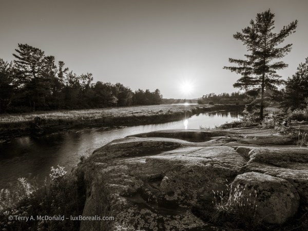 The sun setting on the Chikanishing River with smooth curves of rock in the foreground and a white pine on the right.